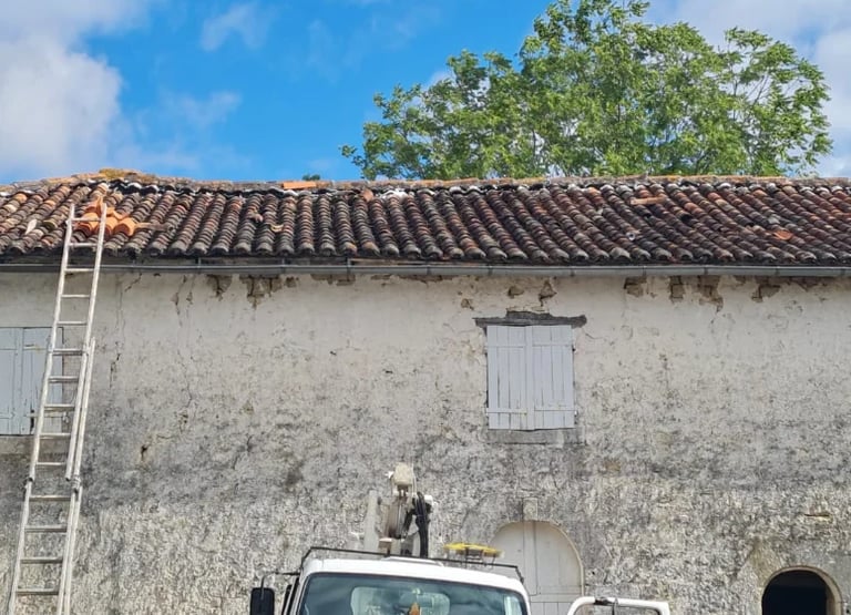 Worker on ladder cleaning weathered white colonial building facade with deteriorating tile roof and vehicles parked below under blue sky