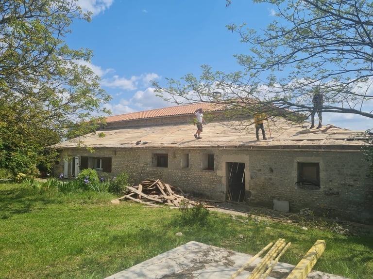 Stone house under renovation with workers on roof, surrounded by green lawn and trees